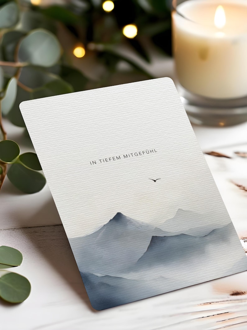 May include: A greeting card with a watercolor mountain landscape in shades of gray and blue. The text "IN TIEFEM MITGEF&Uuml;HL" is printed at the top. A lit candle and greenery are in the background.