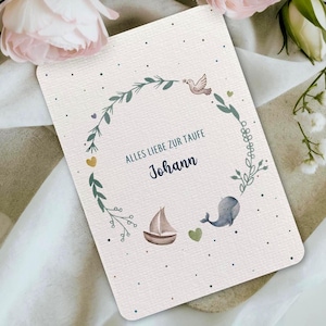 Könnte beinhalten: Eine cremefarbene Karte mit einem Aquarelldesign. Die Karte zeigt einen kreisförmigen Kranz aus grünen Blättern und kleine Illustrationen einer Taube, eines Segelbootes und eines Wals. Der Text "ALLES LIEBE ZUR TAUFE" und der Name "Johann" sind blau gedruckt.