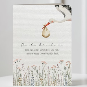 May include: A greeting card with a watercolor illustration of a stork carrying a bundle. The card features text in a script font, with a floral border at the bottom. The card is on a white surface.