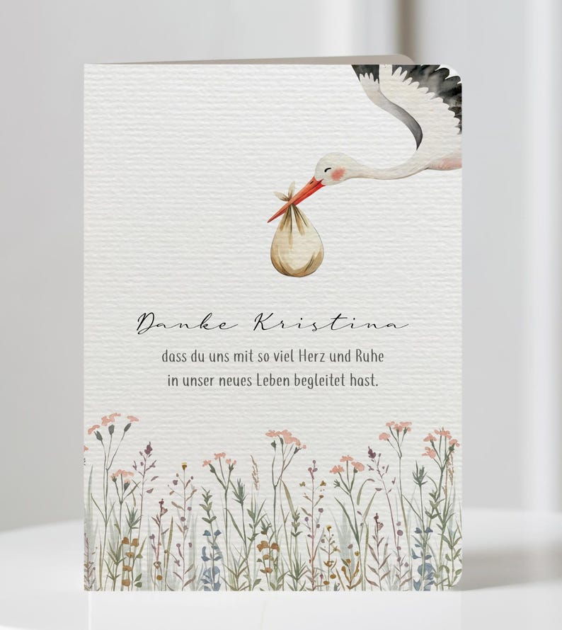 May include: A greeting card with a watercolor illustration of a stork carrying a bundle. The card features text in a script font, with a floral border at the bottom. The card is on a white surface.