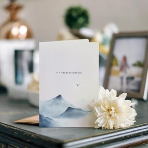 May include: A folded white card with a watercolor painting of blue mountains and the text "IN TIEFEM MITGEF&Uuml;HL". A white flower and a framed photo are next to the card on a dark surface.