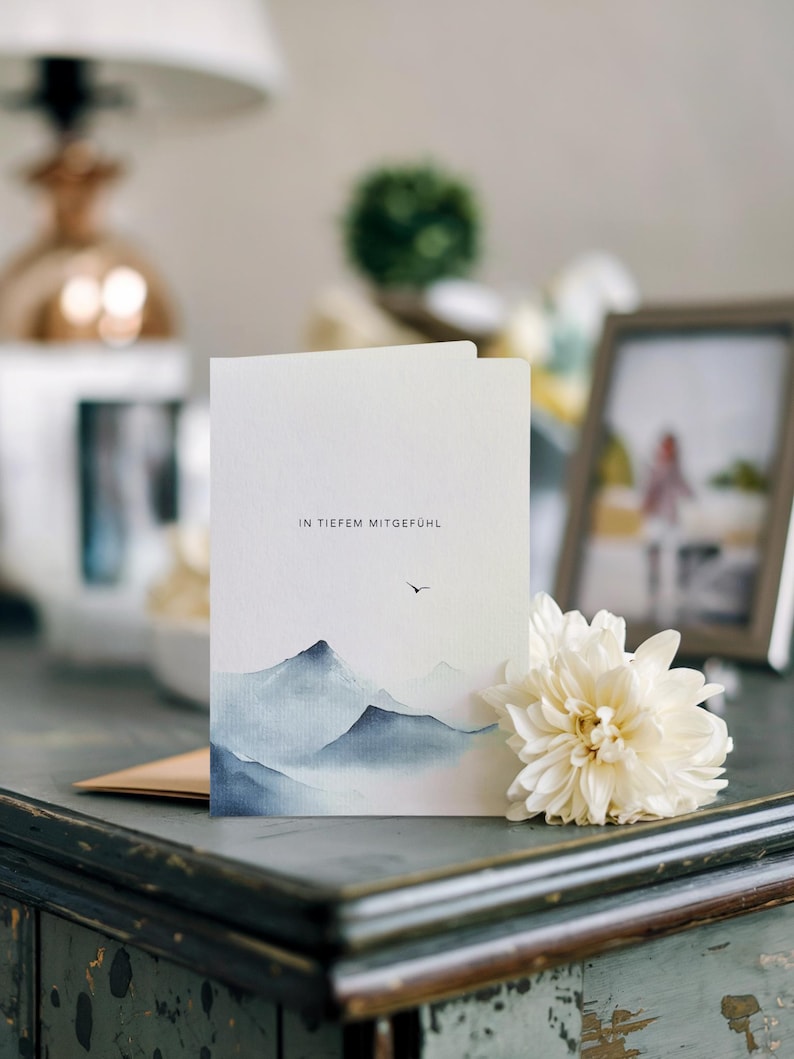 May include: A folded white card with a watercolor painting of blue mountains and the text "IN TIEFEM MITGEF&Uuml;HL". A white flower and a framed photo are next to the card on a dark surface.
