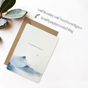 May include: A white greeting card with a watercolor mountain scene in shades of blue and gray, paired with a brown envelope. The card has the text "IN TIEFEM MITGEF&Uuml;HL" and a small bird illustration. The text "wahlweise mit hochwertigem kraftpapierumschlag" is also present.