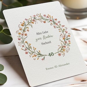 Può includere: Una carta bianca con un design a ghirlanda floreale. La ghirlanda è composta da foglie verdi e piccoli fiori rosa e gialli. Il testo centrale recita "Alles Liebe zur Rubin Hochzeit" e "40". I nomi "Emma & Alexander" sono in basso.