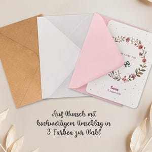 Puede incluir: Una tarjeta de felicitaci&oacute;n con un dise&ntilde;o de corona floral, dentro de un sobre rosa, con un sobre blanco detr&aacute;s y un sobre marr&oacute;n a la izquierda. La tarjeta tiene el nombre "Emma" y la fecha "15. August 2026". El texto "ES LIEBE ZUR" y "fe" tambi&eacute;n son visibles.