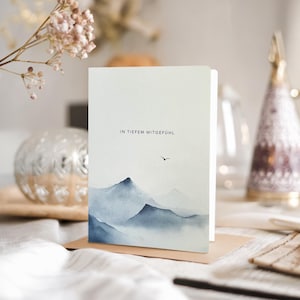 May include: A greeting card with a watercolor mountain landscape in shades of blue and gray. The card features the text "IN TIEFEM MITGEF&Uuml;HL" and a small bird silhouette. The card is displayed on a brown surface.