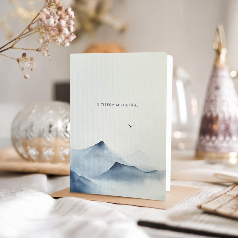 May include: A greeting card with a watercolor mountain landscape in shades of blue and gray. The card features the text "IN TIEFEM MITGEF&Uuml;HL" and a small bird silhouette. The card is displayed on a brown surface.
