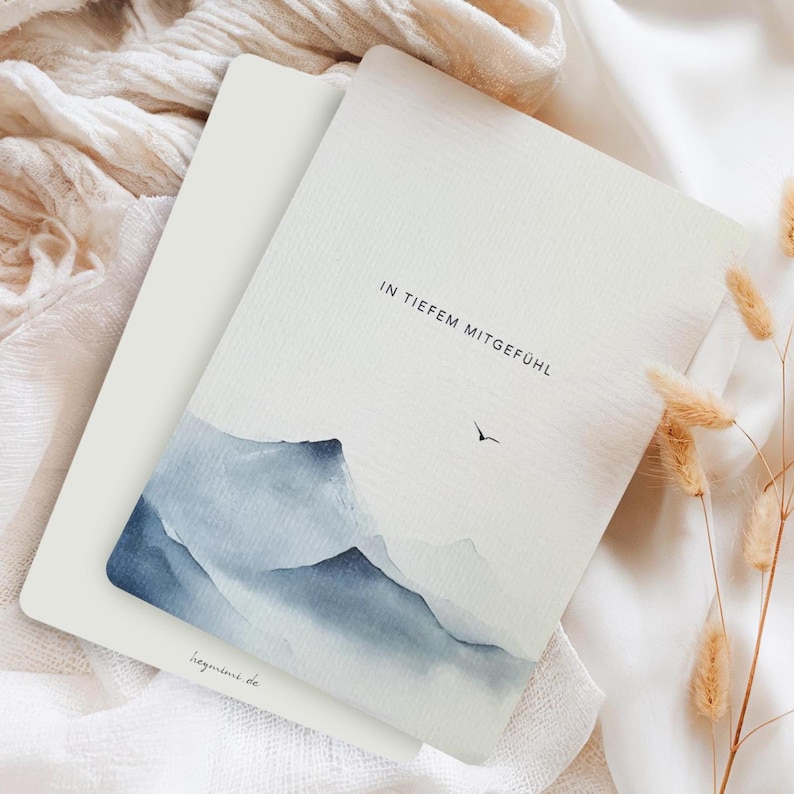 May include: Two white cards with a watercolor mountain scene in shades of blue. The top card has the text "IN TIEFEM MITGEF&Uuml;HL" and a small bird illustration. The bottom card has the text "hegimini.de". The cards are on a white fabric surface.
