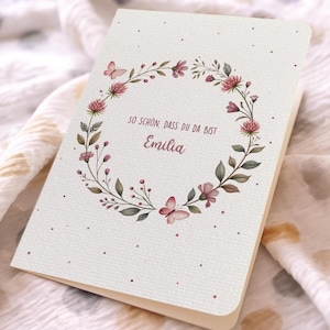 May include: A folded greeting card with a floral wreath design. The wreath features pink flowers, green leaves, and butterflies. The text "SO SCH&Ouml;N, DASS DU DA BIST Emilia" is printed in a script font within the wreath. The card has a cream-colored background.