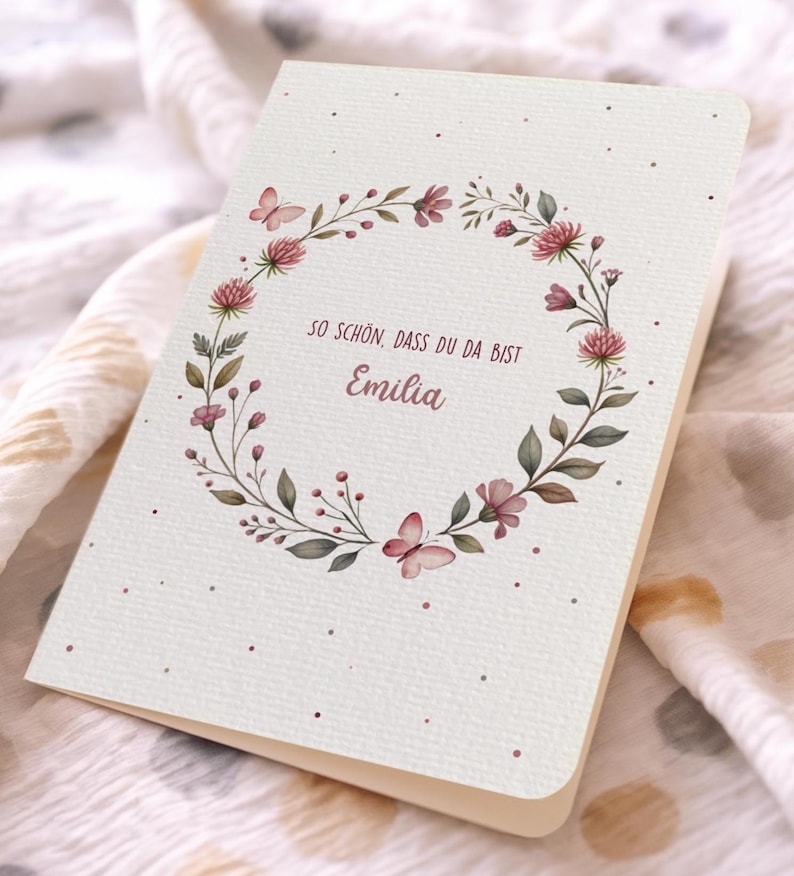 May include: A folded greeting card with a floral wreath design. The wreath features pink flowers, green leaves, and butterflies. The text "SO SCH&Ouml;N, DASS DU DA BIST Emilia" is printed in a script font within the wreath. The card has a cream-colored background.