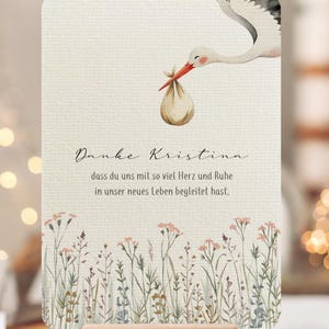 May include: A watercolor-style baby shower card featuring a stork carrying a bundle. The card has text in German and a floral border at the bottom. It is displayed on a wooden block.