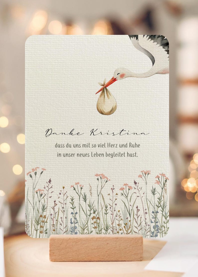 May include: A watercolor-style baby shower card featuring a stork carrying a bundle. The card has text in German and a floral border at the bottom. It is displayed on a wooden block.