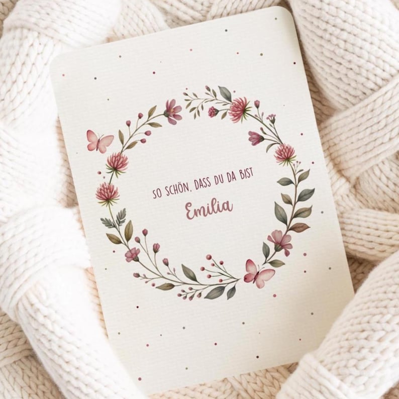May include: A greeting card with a floral wreath design. The wreath is composed of pink flowers, green leaves, and small butterflies. The card has the name "Emilia" and the text "SO SCH&Ouml;N, DASS DU DA BIST".