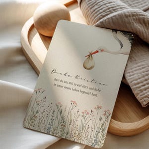 May include: A greeting card with a watercolor illustration of a stork carrying a bundle. The card has text in a script font and a floral border at the bottom. The card is on a wooden tray.