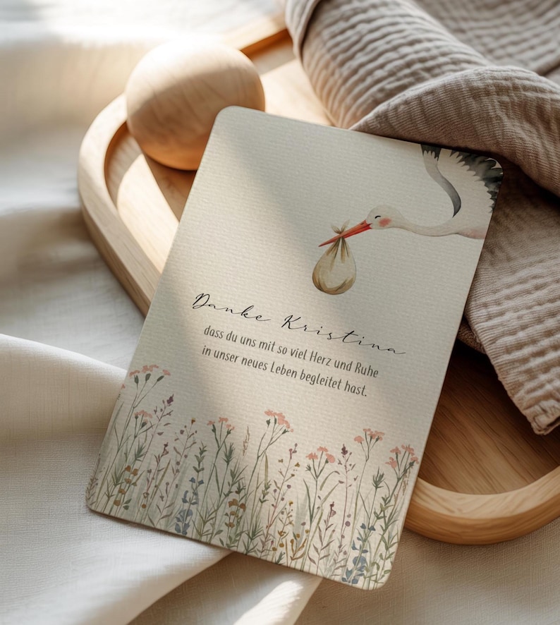 May include: A greeting card with a watercolor illustration of a stork carrying a bundle. The card has text in a script font and a floral border at the bottom. The card is on a wooden tray.