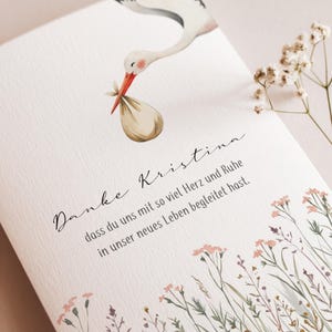 May include: A greeting card with a watercolor illustration of a stork carrying a bundle. The card has text in script and a floral border. The stork is white, black, and orange.