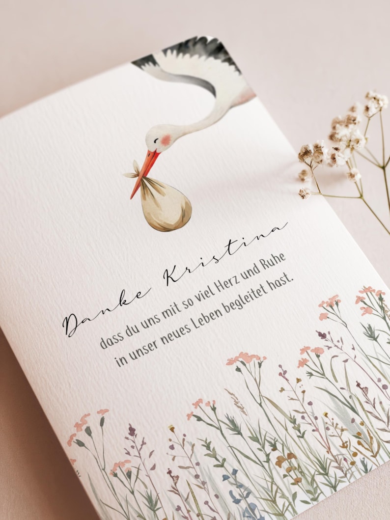 May include: A greeting card with a watercolor illustration of a stork carrying a bundle. The card has text in script and a floral border. The stork is white, black, and orange.