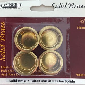 May include: Four solid brass flush pulls, each with a circular design. The package includes the text "Solid Brass" and "3/4" 19mm". The brand name "BRAINERD" is visible at the top. The package also includes the text "4 Flush Fi", "4 Poignées", and "4 Asas Para D".