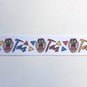 May include: White ribbon with a repeating pattern of Taz the Tasmanian Devil's face, the word "Taz" and colorful triangles.