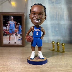 Puede incluir: Figura de bobblehead de un jugador de baloncesto con una camiseta azul con la palabra "Thunder" y el número 2. La figura sostiene una pelota de baloncesto y está sobre una base de madera. Una foto enmarcada y trofeos en el fondo.