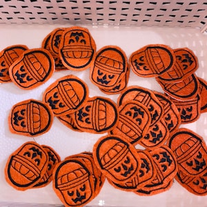 May include: A collection of orange felt Halloween basket patches with black embroidered details. The patches feature a jack-o'-lantern face and basket design. The patches are in a white plastic basket.