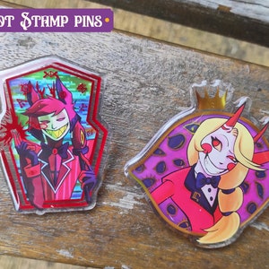Peut inclure: Deux &eacute;pingles en acrylique repr&eacute;sentant des personnages de la s&eacute;rie anim&eacute;e "Hazbin Hotel". La premi&egrave;re &eacute;pingle est une &eacute;pingle en forme de cercueil rouge et noir avec une image de dessin anim&eacute; d'un d&eacute;mon aux cheveux roux avec des cornes et un n&oelig;ud papillon. La deuxi&egrave;me &eacute;pingle est une &eacute;pingle ronde avec une image de dessin anim&eacute; d'un d&eacute;mon blond avec des cornes et un n&oelig;ud papillon. L'arri&egrave;re-plan de la deuxi&egrave;me &eacute;pingle est violet avec des yeux dor&eacute;s.