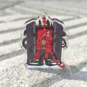 May include: A clear acrylic charm featuring a character in a red coat, standing before a dark portal. Black shadowy figures surround the portal. A red beaded chain hangs from the top of the charm.
