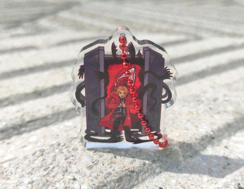 May include: A clear acrylic charm featuring a character in a red coat, standing before a dark portal. Black shadowy figures surround the portal. A red beaded chain hangs from the top of the charm.