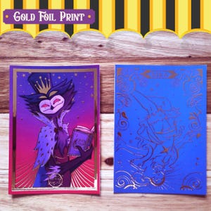 May include: Two gold foil printed cards. The first card features a purple and pink background with a black and white cartoon character wearing a crown and holding a book. The second card features a blue background with a gold foil cartoon character with a star on their chest. The text "MIKA" is printed on the card.