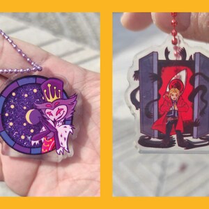 May include: Two acrylic keychains. One features a purple and white cartoon character with a crown, set against a starry night background. The other depicts a character in a red coat in front of a red portal.