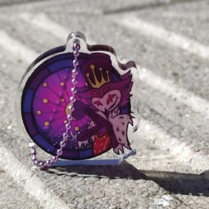 May include: A clear acrylic keychain featuring a cartoon owl character with a crown, set against a stained-glass window design in purple and blue. The keychain has a silver chain and a small base.