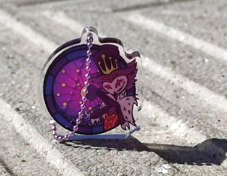 May include: A clear acrylic keychain featuring a cartoon owl character with a crown, set against a stained-glass window design in purple and blue. The keychain has a silver chain and a small base.