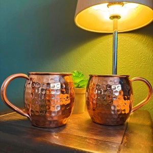 Puede incluir: Dos jarras de cobre con textura martillada. Las jarras tienen una forma redondeada y un asa en el lateral. Las jarras están sobre una superficie de madera.