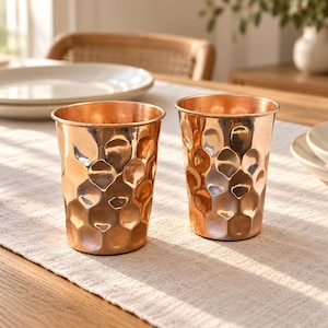 May include: Two matching copper tumblers featuring a distinct hammered texture across the exterior surface. Each cup has a cylindrical shape with a smooth rim and a polished metallic finish that reflects light, creating a warm, reflective appearance.