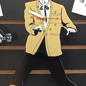 Pode incluir: Relógio de parede com uma imagem estilizada de Elvis Presley com um casaco dourado e calças pretas, numa pose. O relógio tem um mostrador branco com números e ponteiros pretos, e um ponteiro de segundos vermelho. O relógio está montado num fundo preto.