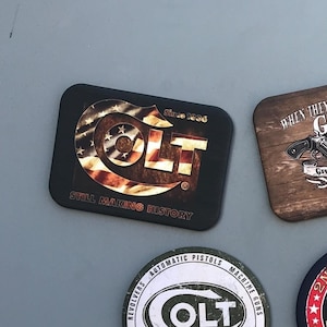 May include: A collection of ten gun-themed refrigerator magnets. The magnets feature various designs, including a skull with crossed pistols, a warning sign that says "This door is locked for your protection, not mine", and a welcome sign that says "Now smile and wait for the flash!"