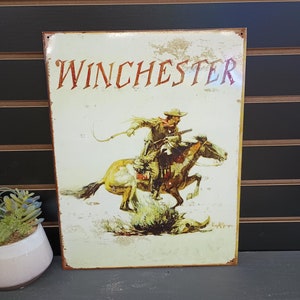 May include: A vintage-style metal sign featuring a cowboy riding a horse. The sign is cream-colored with brown and red accents. The text "Winchester" is printed in red at the top of the sign.