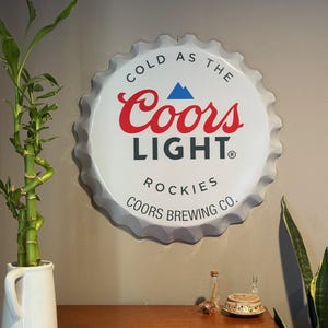 Könnte beinhalten: Weiße Metall-Wanddekoration in Form eines Coors Light Bierflaschenverschlusses mit dem Text "Cold as the Coors Light" und "Rockies Coors Brewing Co."