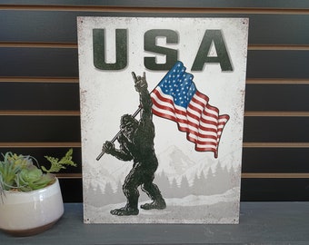 Puede incluir: Un letrero de metal con un fondo blanco desgastado que presenta un Bigfoot de dibujos animados sosteniendo una bandera estadounidense con la palabra "USA" en letras verdes sobre el Bigfoot.