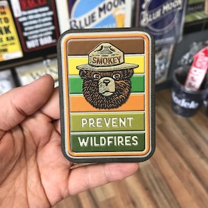 May include: A brown and green patch with a cartoon bear wearing a ranger hat. The hat says "Smokey" and the patch says "Prevent Wildfires".