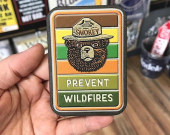 May include: A brown and green patch with a cartoon bear wearing a ranger hat. The hat says "Smokey" and the patch says "Prevent Wildfires".