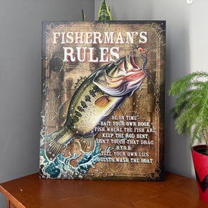 以下が含まれることがあります： 「FISHERMAN'S RULES」というテキストと釣りに関するルールのリストが書かれた、素朴なスタイルの看板。看板には、水面から飛び出すブラックバスが描かれている。背景は、使い古した木の質感である。