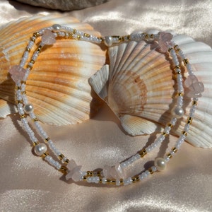 Op de afbeelding: Een delicate ketting met twee rijen witte en gouden kralen, versierd met roze bloemvormige bedels. De ketting ligt op een witte stoffen achtergrond met schelpen.