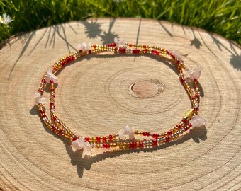 May include: A beaded waist chain with red and gold beads and pink stone accents. The chain is lying on a wooden surface.