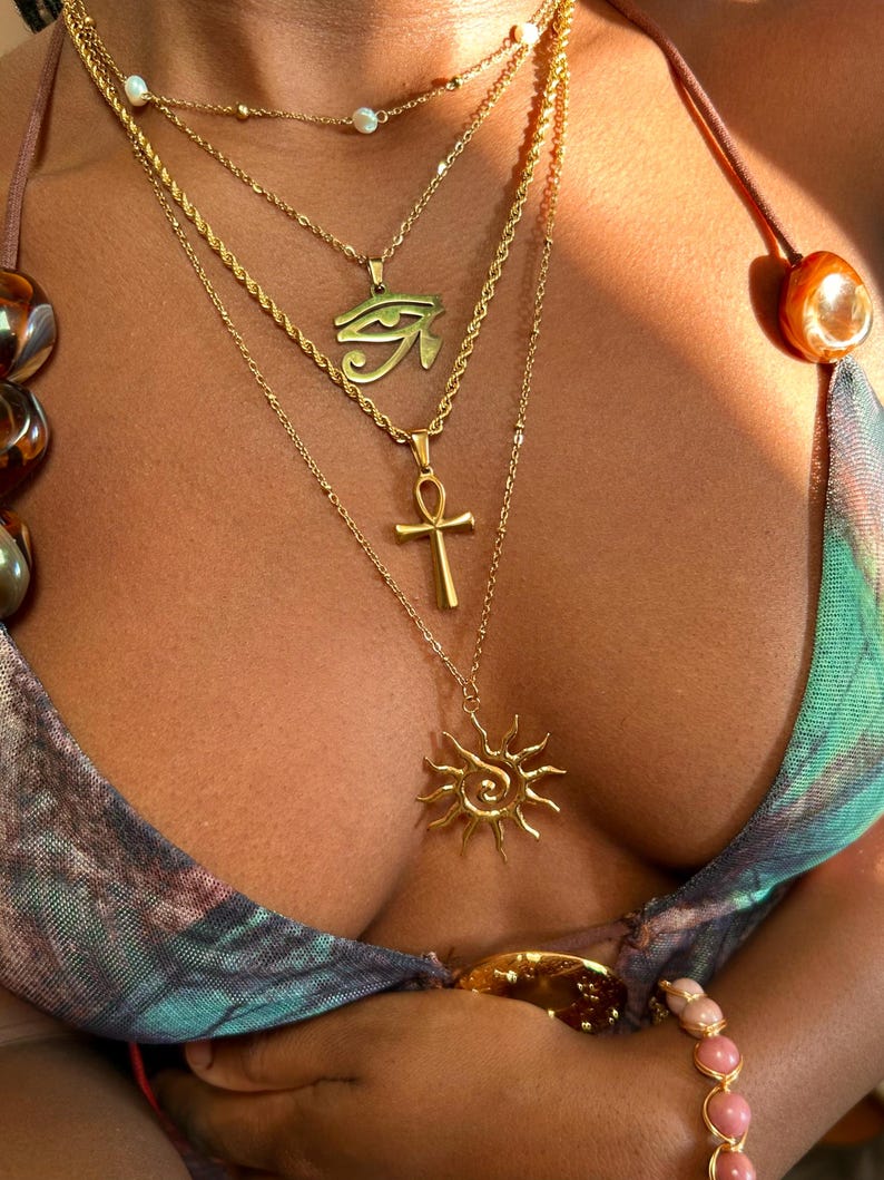 Peut inclure: Plusieurs colliers dor&eacute;s avec diff&eacute;rents pendentifs, dont un &OElig;il d'Horus, une croix Ankh et un soleil. Les colliers sont superpos&eacute;s et port&eacute;s avec un haut &agrave; motifs et un bracelet de perles roses.