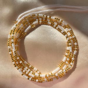 Op de afbeelding: Waist beads met afwisselend gouden en witte kralen. De kralen zijn geregen op wit draad en in een cirkelvormig patroon gerangschikt. De achtergrond is een lichte, satijnachtige stof.