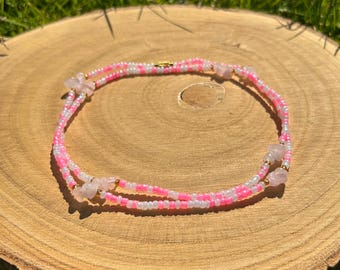 May include: A delicate beaded body chain in pink and white, accented with small rose quartz stones. The jewelry is displayed on a wooden surface, with green grass in the background. The chain is designed to be worn around the waist.