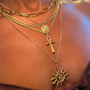 Peut inclure: Plusieurs colliers dor&eacute;s superpos&eacute;s. Les colliers comprennent une cha&icirc;ne &eacute;paisse, une cha&icirc;ne torsad&eacute;e, un pendentif avec un motif circulaire, un pendentif ankh et un pendentif soleil. Les bijoux sont port&eacute;s sur un haut marron.
