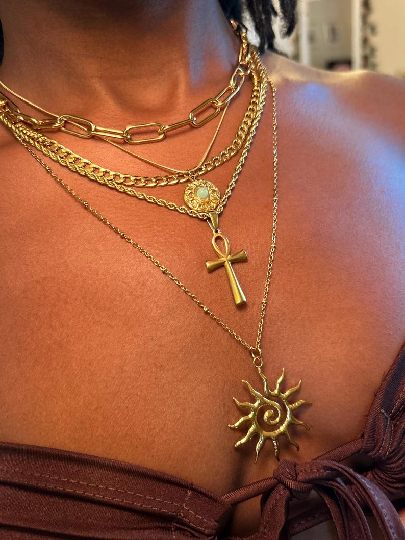 Peut inclure: Plusieurs colliers dor&eacute;s superpos&eacute;s. Les colliers comprennent une cha&icirc;ne &eacute;paisse, une cha&icirc;ne torsad&eacute;e, un pendentif avec un motif circulaire, un pendentif ankh et un pendentif soleil. Les bijoux sont port&eacute;s sur un haut marron.