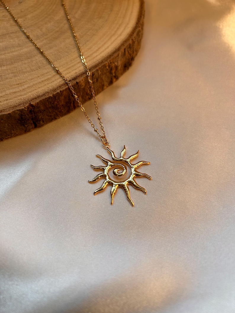 Peut inclure: Un collier dor&eacute; avec un pendentif soleil. Le pendentif pr&eacute;sente un centre en spirale avec des rayons rayonnants. La cha&icirc;ne du collier est d&eacute;licate et le pendentif soleil est centr&eacute;. Le fond est un tissu de couleur claire.